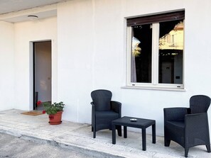 A clean, white exterior wall with a large window, two wicker chairs, a small table, and a potted plant.
