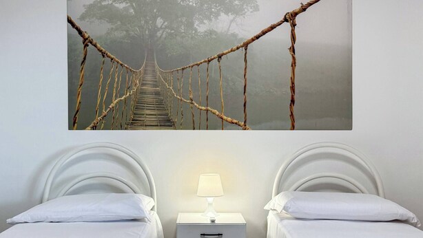 Una habitació amb dos llits, una tauleta de nit amb una làmpada, i una imatge gran d'un pont de corda al fons.