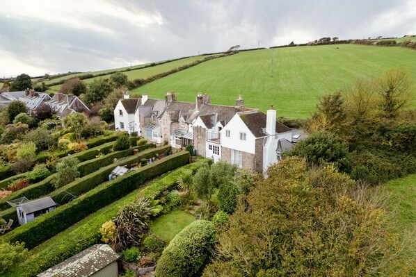 A large, white, two-story house with a well-maintained garden and a green field in the background. - Pear Tree Cottage in South Milton (Kingsbridge)