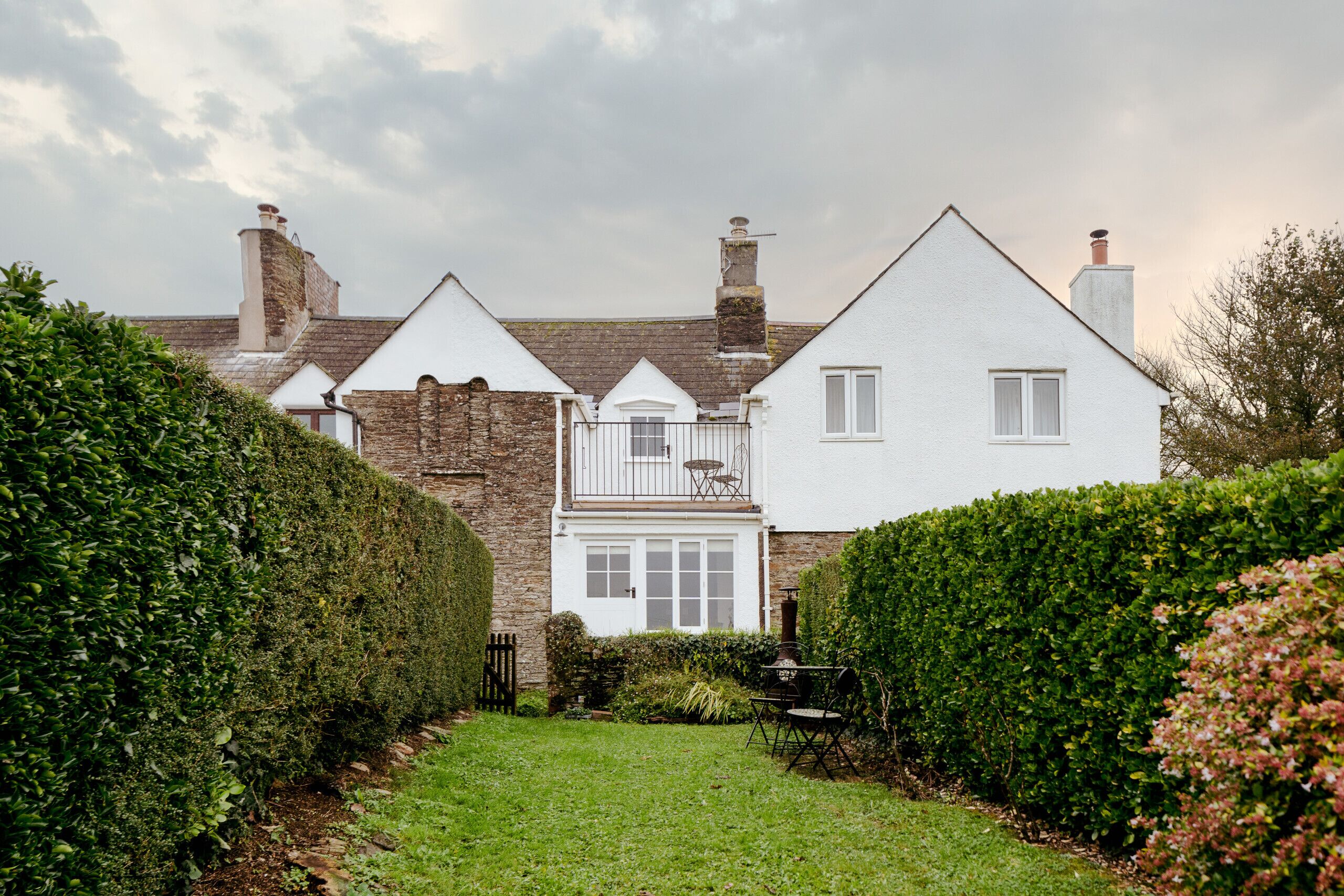 Une maison blanche traditionnelle, dotée d’une cheminée en pierre, d’un petit balcon et d’un jardin bien entretenu avec des haies et une pelouse.