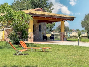 A covered outdoor seating area with two chairs and a table, surrounded by greenery and a stone building.