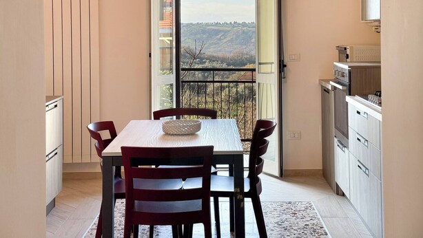A dining area with a table and chairs, a view through a glass door, and a kitchen with appliances.