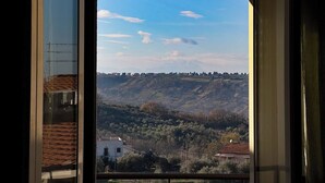 A view through a window of a building with a balcony, overlooking a hilly landscape with scattered houses and trees.