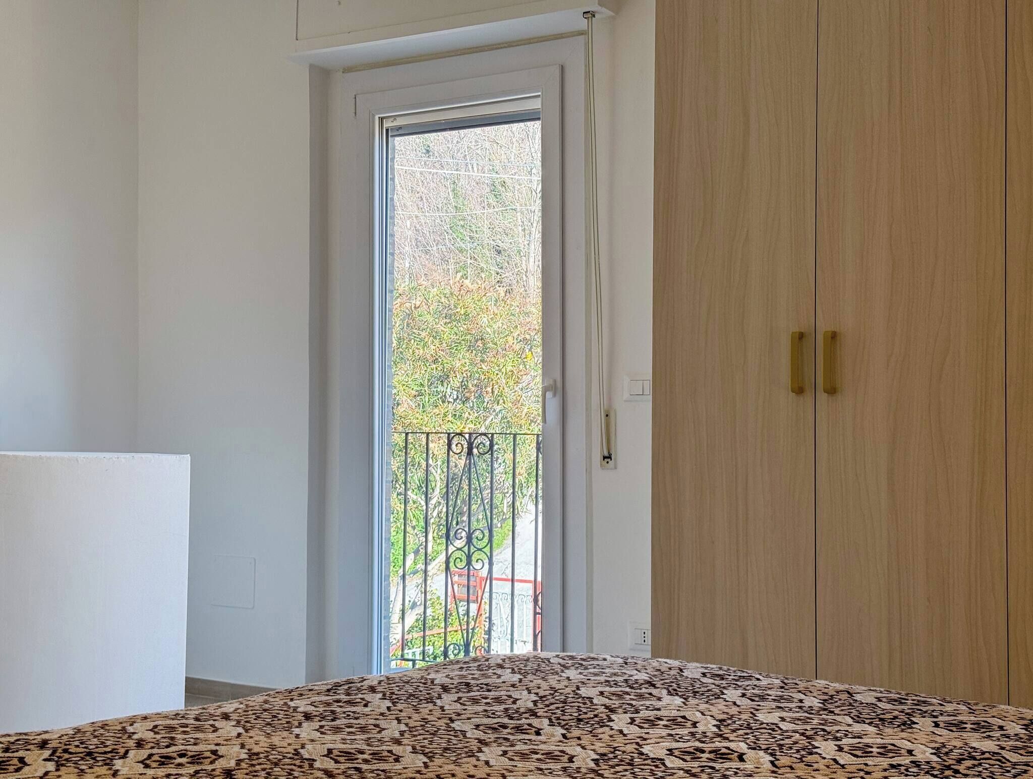 A room with a bed, a wardrobe, a door leading to a balcony with a metal railing, and a view of greenery outside.