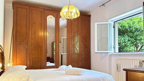 Quarto com cama, guarda-roupa de madeira, espelho, janela com vista para o verde e lustre.