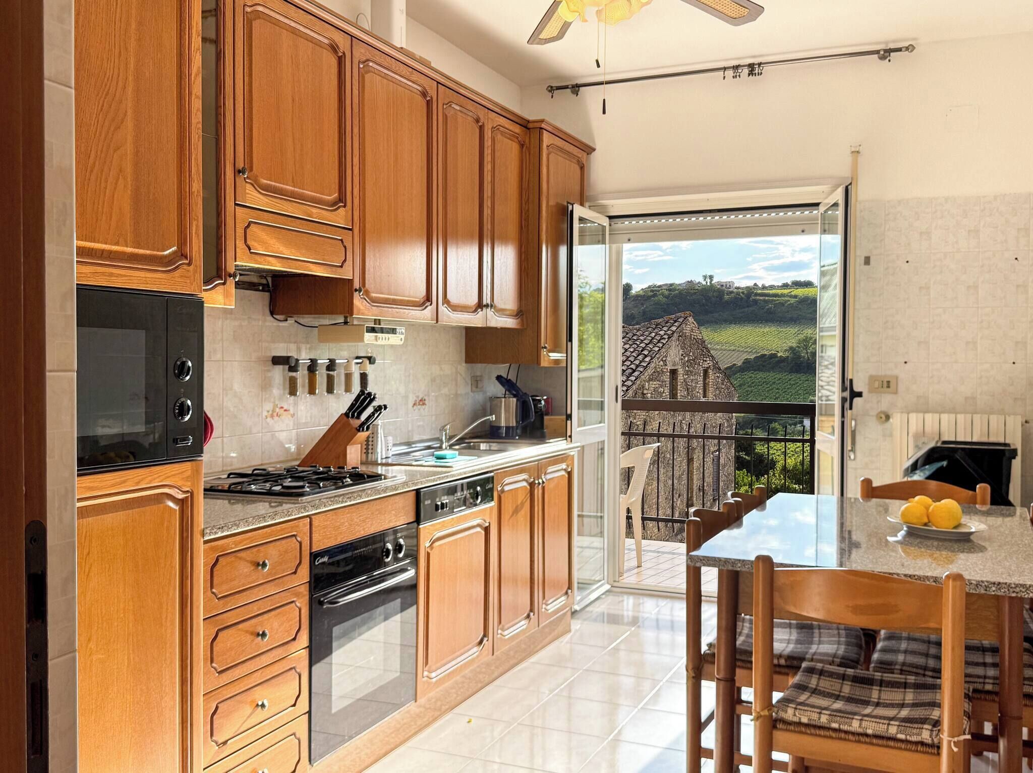 A kitchen with wooden cabinets, a stove, and a dishwasher. There is a dining area with a table and chairs, and a view of a vineyard through a glass door.