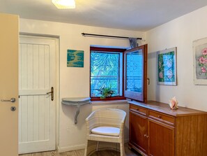 A room with a white door, a window with a view, a wicker chair, a wooden cabinet, and wall art.
