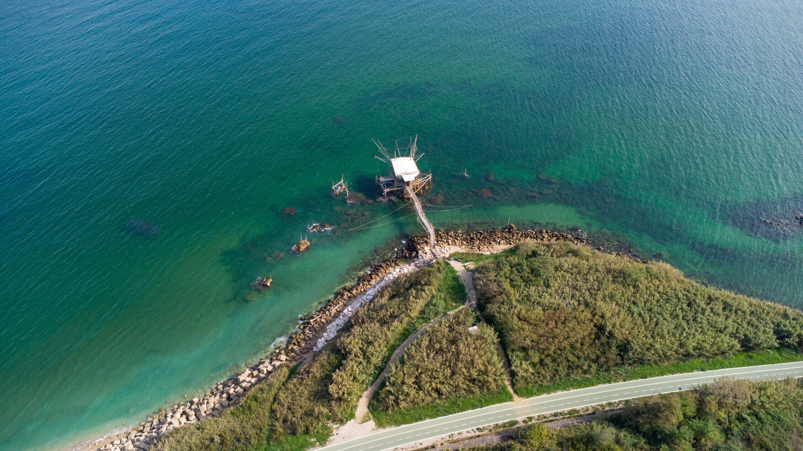An aerial view of a coastal area with a wooden structure on stilts, a rocky shoreline, a paved road, and dense greenery.