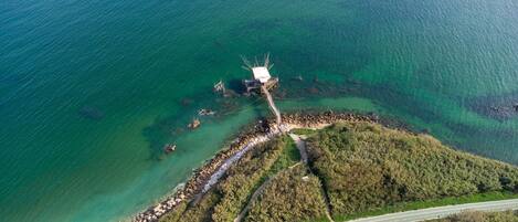 An aerial view of a coastal area with a wooden structure on stilts, a rocky shoreline, a paved road, and dense greenery.