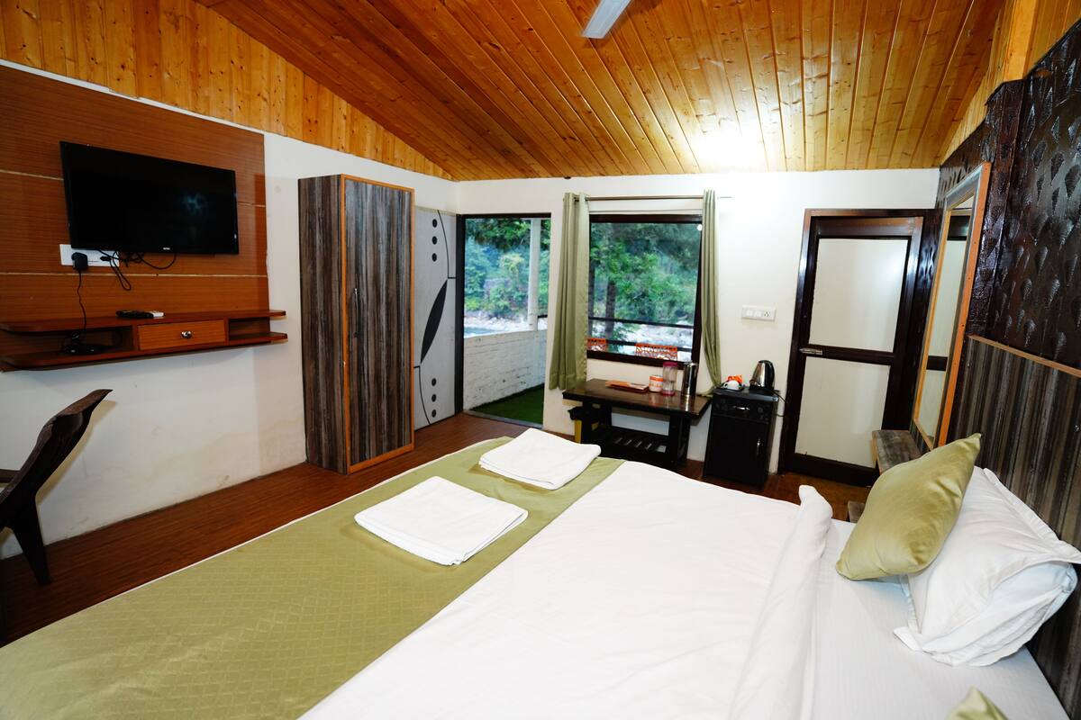 A hotel room with a wooden ceiling, a bed with white linens, a TV mounted on the wall, and a window with curtains.