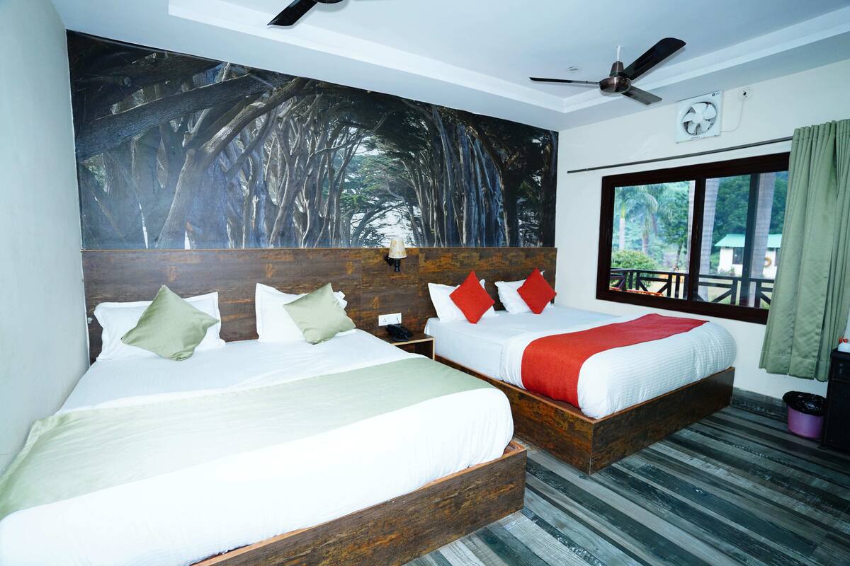 A hotel room with two beds, a large forest mural, a ceiling fan, a window with a view, and a wooden floor.