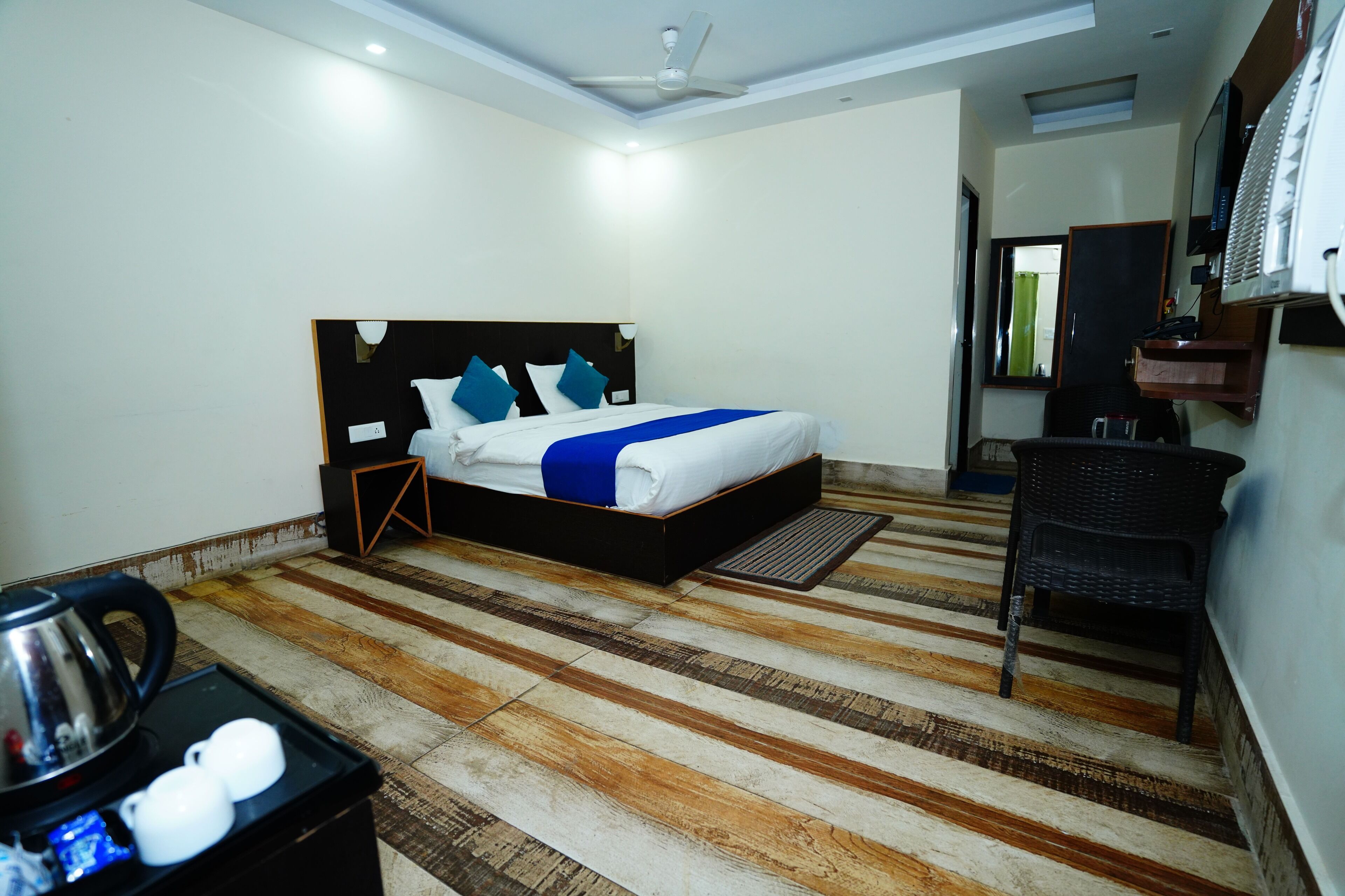 A hotel room with a bed, a chair, a desk with a kettle, and a small table with cups.