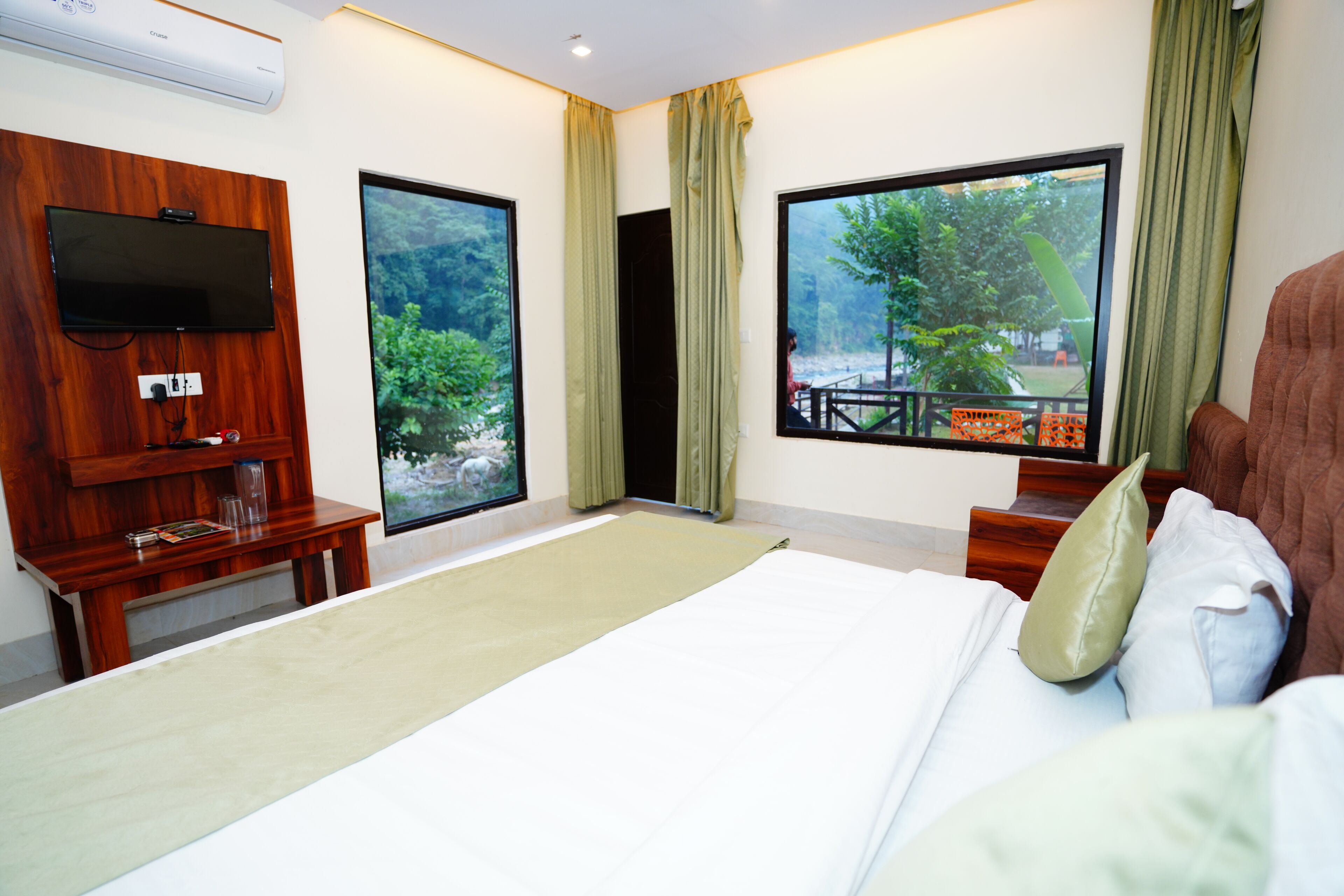 Habitación de hotel con una cama grande, un televisor montado en la pared y dos ventanas con cortinas.