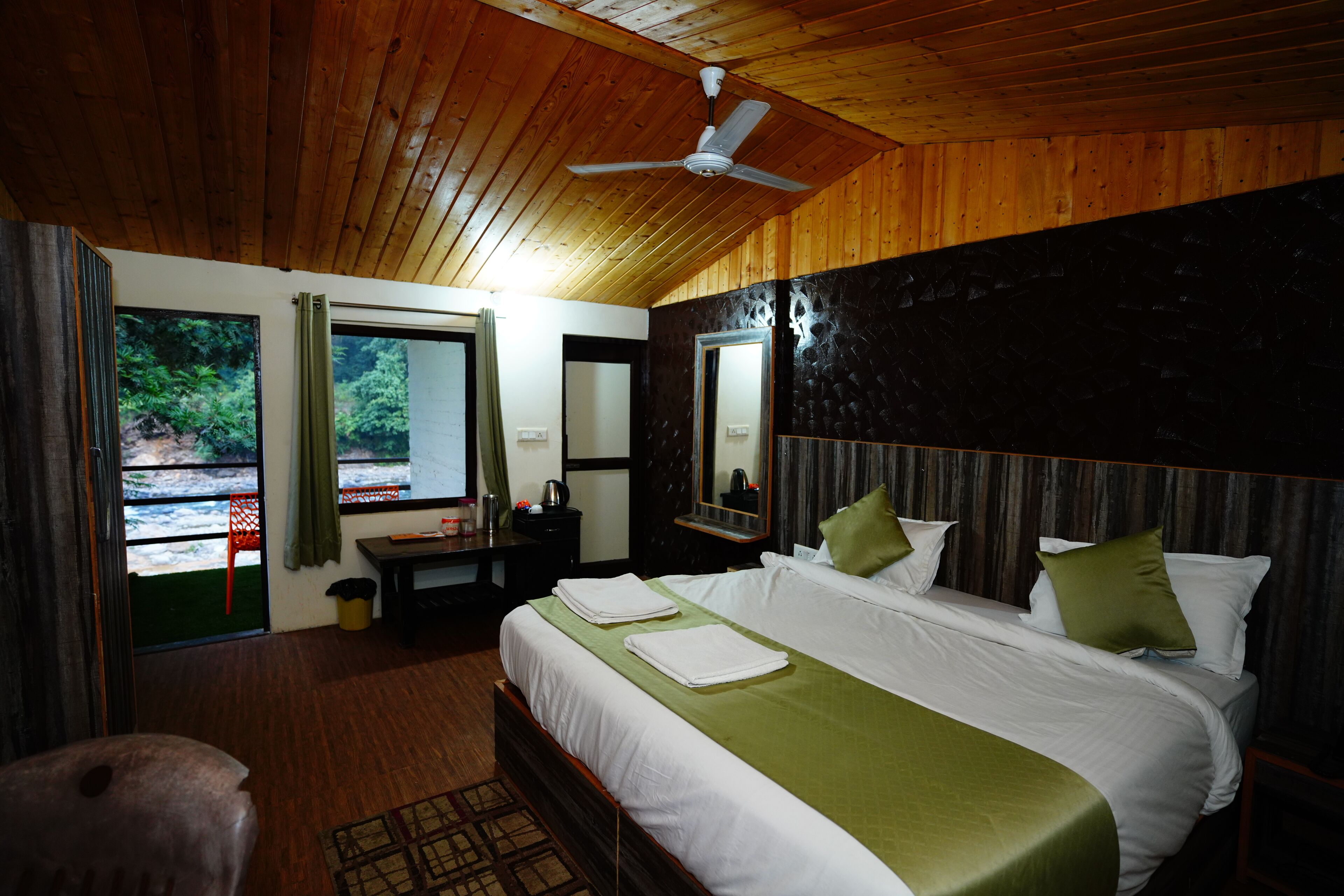 A hotel room with a large bed, a ceiling fan, and a view of greenery outside.