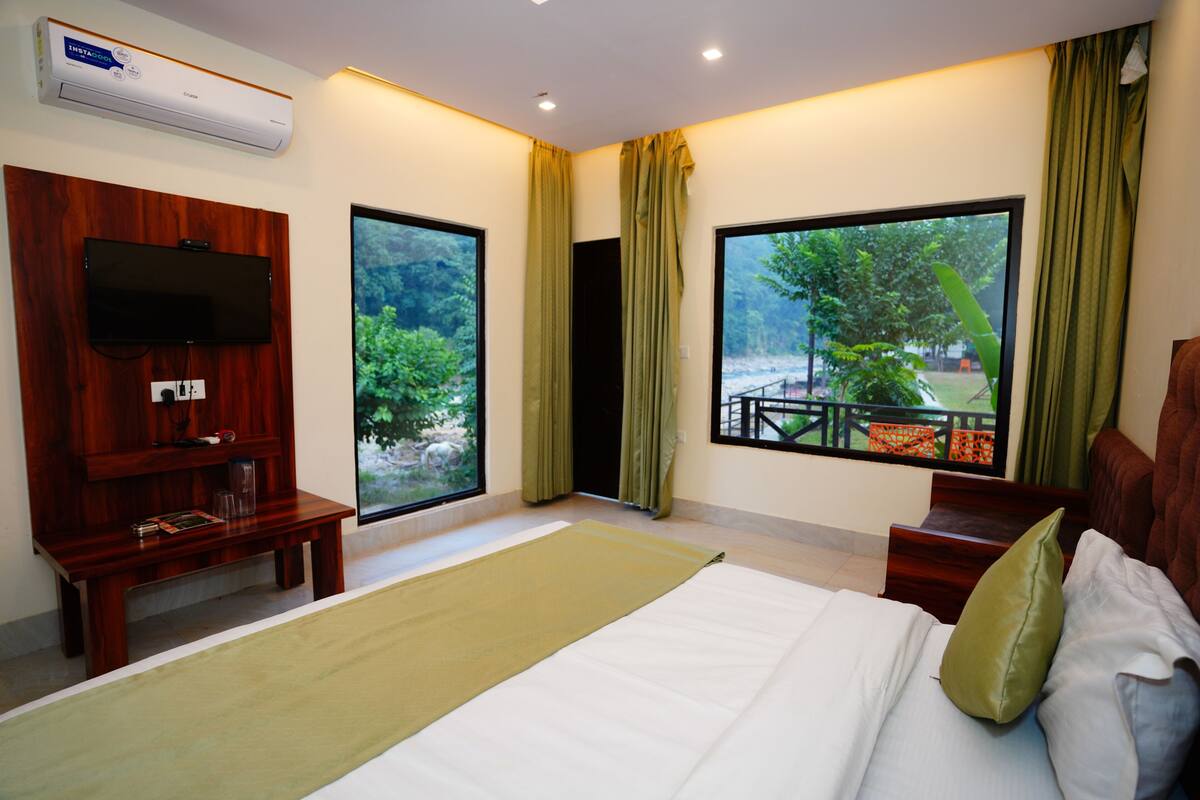 A hotel room with a large bed, a flat-screen TV mounted on a wooden panel, a small wooden bench, and large windows with curtains.