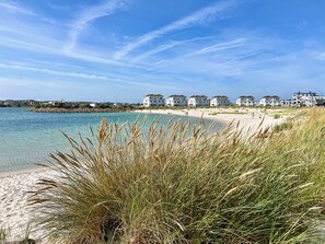 Une plage à l’eau claire, bordée de dunes verdoyantes, avec des bâtiments blancs en arrière-plan.