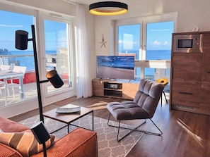 Een moderne woonkamer met een grote televisie, comfortabele zitplaatsen, een houten kast en uitzicht op zee.