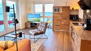 A modern living room with a large TV, comfortable seating, a kitchen area with a microwave, and a view of the sea through large windows. - Penthouse Olpenitz Living - Sea View, Sauna, Elevator, Underground Parking Space (Olpenitz)