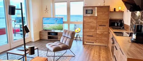 Un salon moderne doté d’un grand téléviseur, de sièges confortables, d’un coin cuisine avec un four à micro-ondes et offrant une vue sur la mer grâce à de grandes fenêtres.