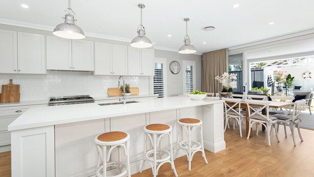 A modern kitchen with white cabinetry, a central island with stools, pendant lighting, and a dining area with a view. - Eastport - Luxury Beach Retreat with Plunge Pool (Catherine Hill Bay)
