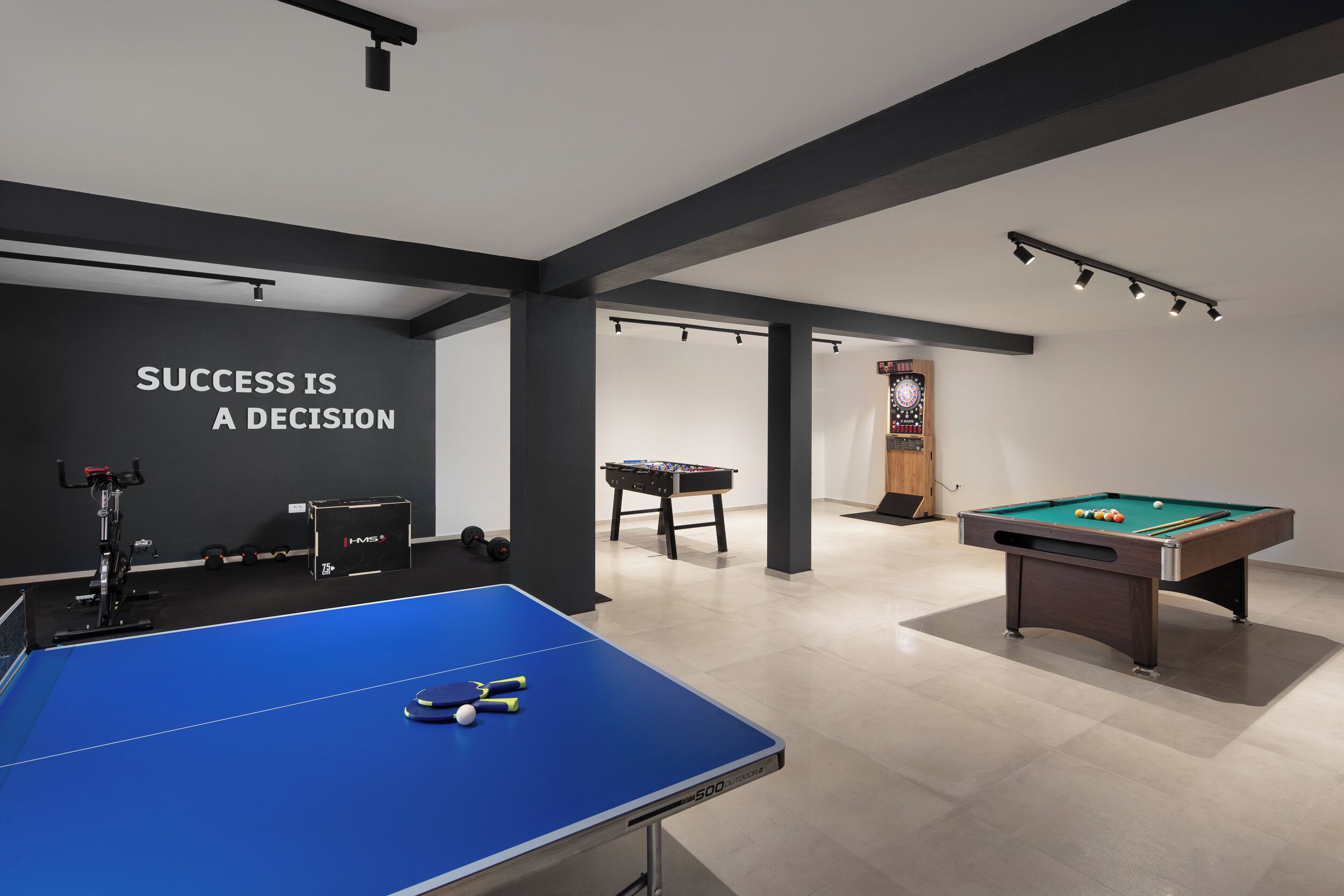 A room with a blue ping pong table, a pool table, and a foosball table. The wall has a motivational quote.