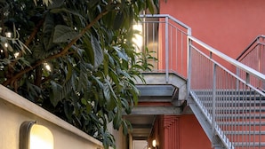 Um corredor com escada, parede com luminárias e fachada de um prédio vermelho.