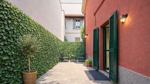 Um beco estreito com uma sebe verde de um lado, uma cesta de vime com uma planta em vaso e um edifício com persianas verdes e um candeeiro.