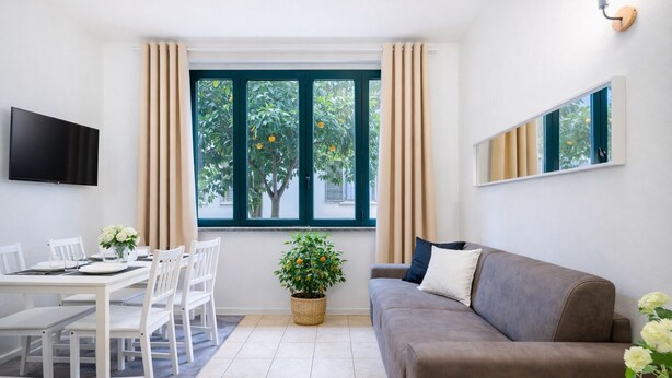 Uma sala de estar moderna com área de jantar, um sofá cinzento, televisão na parede e uma janela com cortinas.
