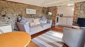 Un salon chaleureux avec un mur en pierre, un canapé gris, une table à manger en bois et un coin cuisine doté de meubles blancs.