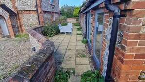 Un étroit sentier mène à un patio aménagé avec une table et des chaises, le tout étant entouré de murs en briques et d’un bâtiment en pierre.