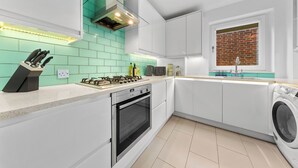 A modern kitchen with white cabinets, a gas stove, a built-in oven, a washing machine, and a window with a brick wall view.