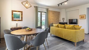 A modern living room with a round wooden dining table, grey upholstered chairs, a yellow sofa, a stone accent wall, a flat-screen TV, and a large window with curtains.