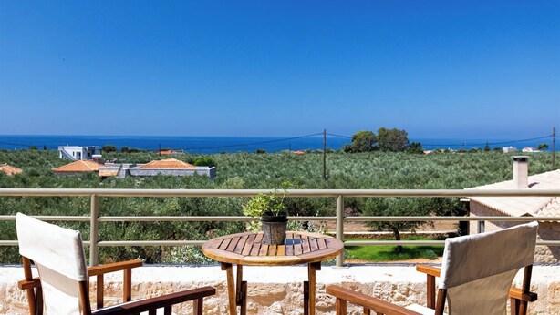A terrace with a round wooden table and two chairs, overlooking a coastal landscape with buildings and trees.