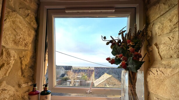 Una ventana con vista a un muro de piedra y un cielo despejado. En el interior, un jarrón con flores secas y dos botellas sobre un repisa.