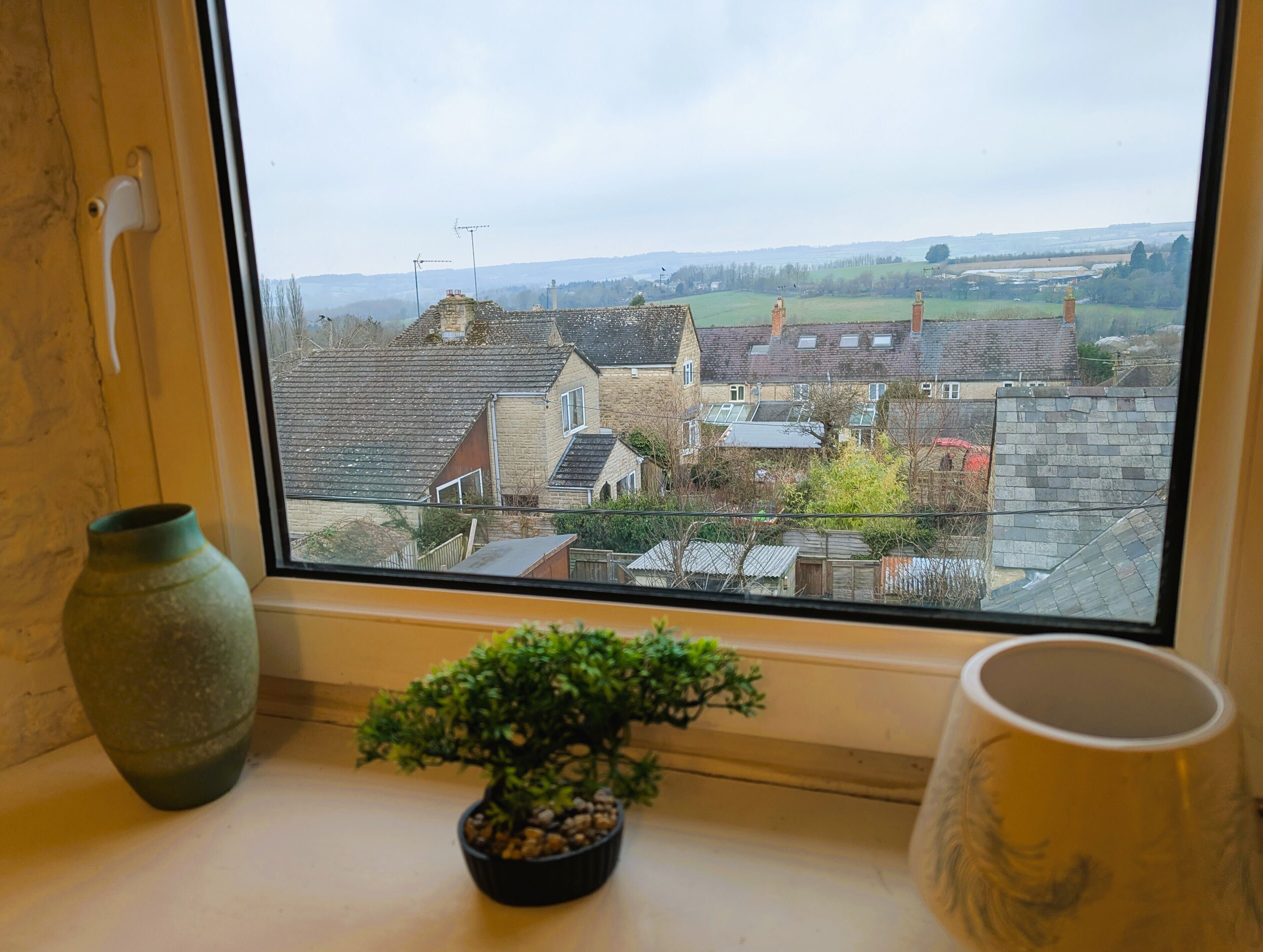Une fenêtre donnant sur un village, une plante en pot et un vase posés sur le rebord de la fenêtre.