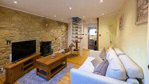 Una sala de estar con pared de piedra, mueble de entretenimiento de madera, televisor de pantalla plana, mesa de centro de madera, sofá blanco y una escalera que sube a un nivel superior.