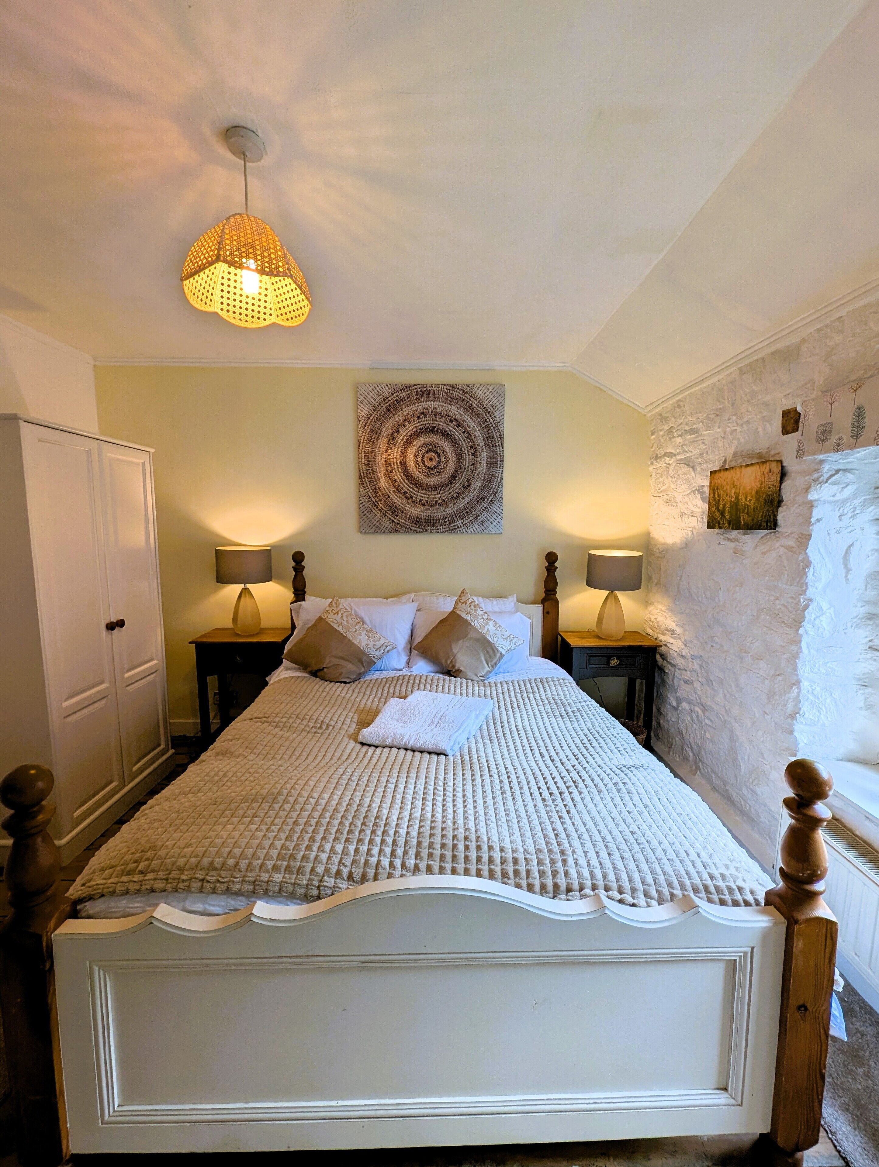 A neatly made bed with a white headboard, two bedside lamps, a wall-mounted circular artwork, and a rustic stone wall.