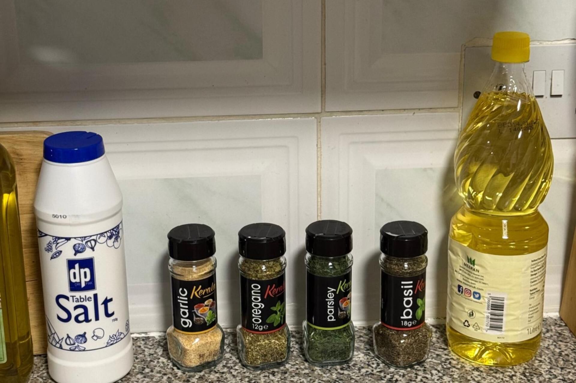 En køkkenbordplade med forskellige krydderier, heriblandt en flaske bordsalt, hvidløg, oregano, persille og basilikum.