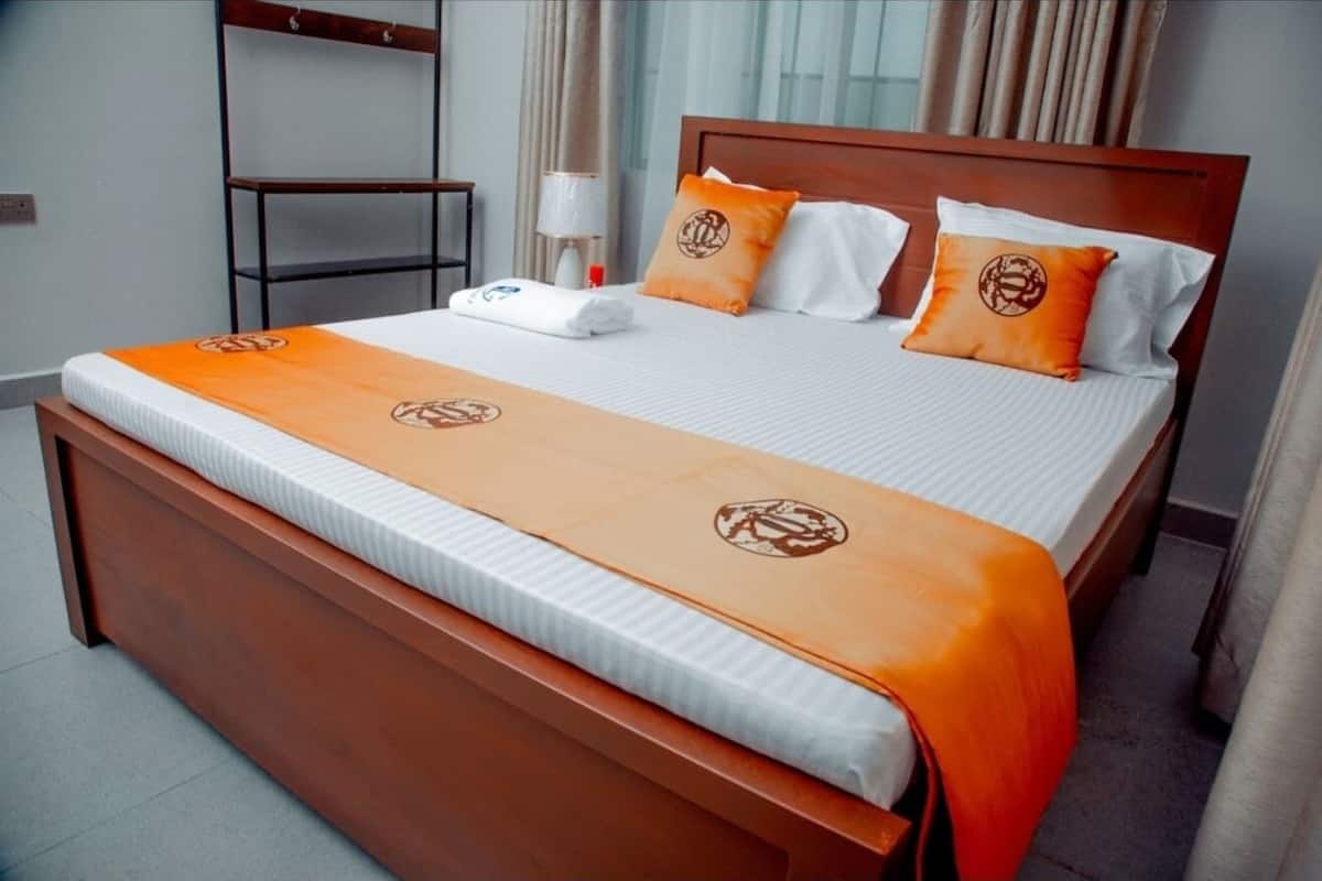 Habitación de hotel con cama de madera, ropa de cama blanca y almohadillas naranjas con un logotipo circular.