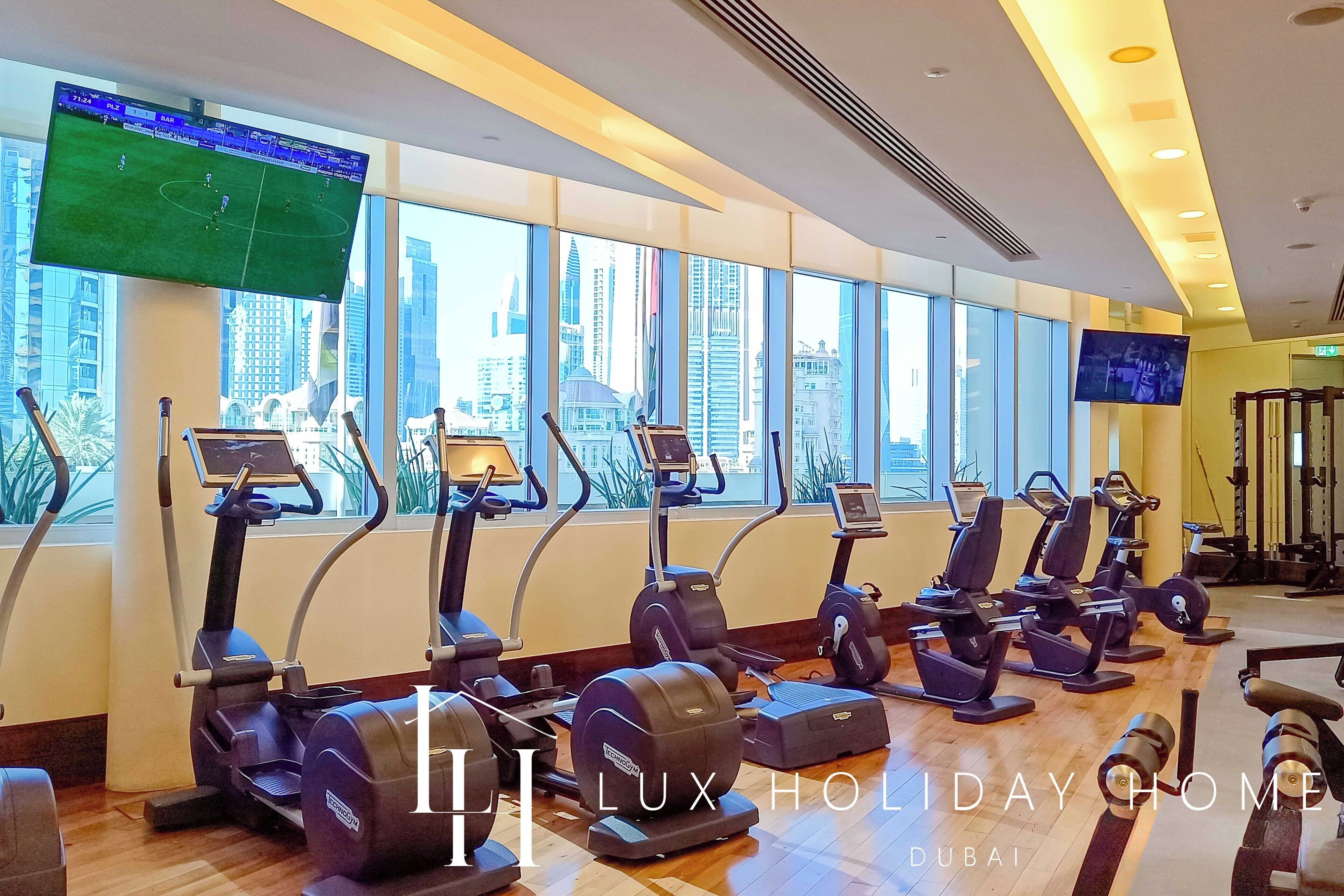 A gym with multiple treadmills and exercise bikes, large windows with a city view, and a television screen displaying a sports game.