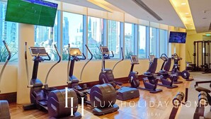 A gym with multiple treadmills and exercise bikes, large windows with a city view, and a television screen displaying a sports game.