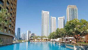 A swimming pool with lounge chairs and umbrellas, surrounded by trees and buildings, including a tall EMAAR building.