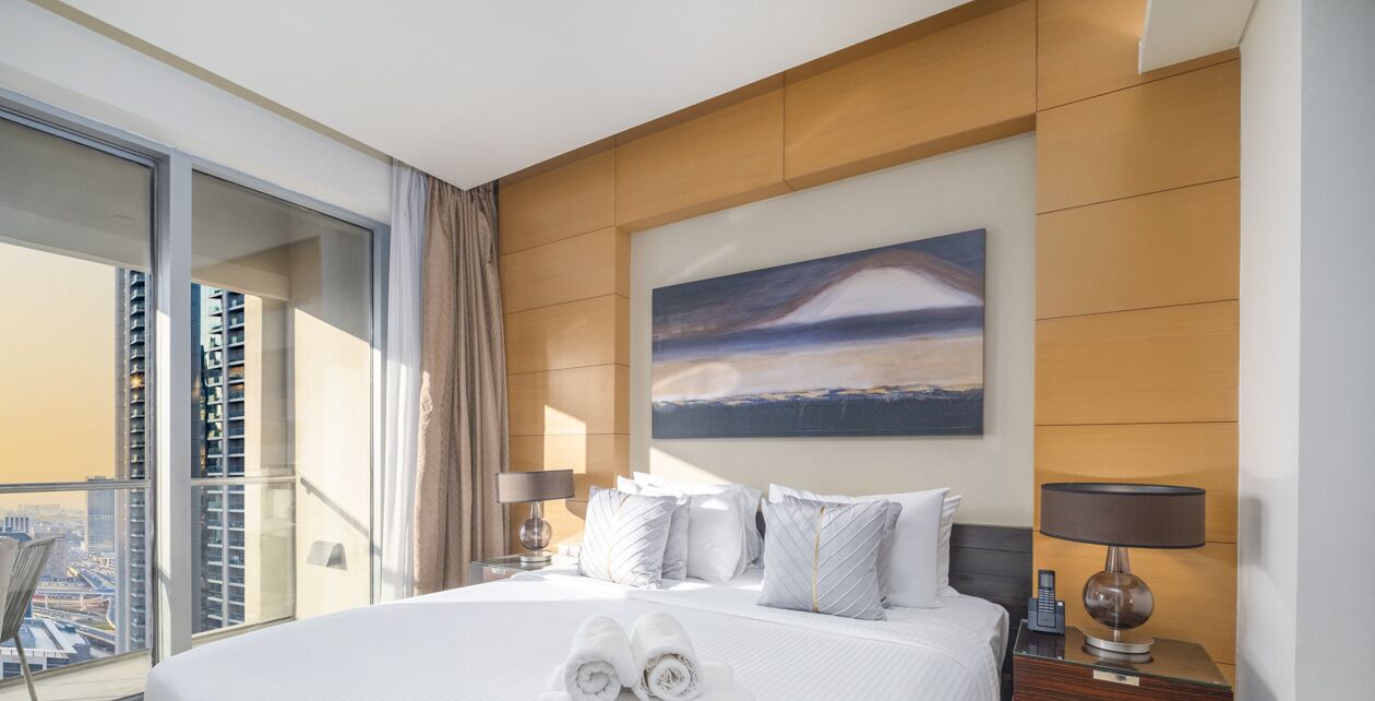 Hotelkamer met een groot bed, een schilderij van een landschap, uitzicht op een stadsbeeld via een balkon, en een net opgemaakt bed met handdoeken.