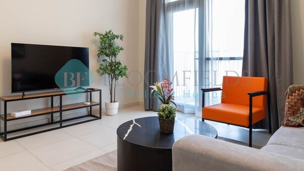 Een moderne woonkamer met een flatscreen televisie, een ronde salontafel, een oranje fauteuil, een grijze bank en een groot raam met gordijnen.
