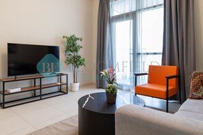 A modern living room with a flat-screen TV, a round coffee table, an orange chair, a grey sofa, and a large window with curtains. - Bloomfields Elegant 1BR in Reflections Al Reem (Abu Dabi)