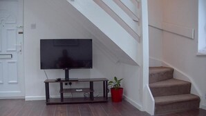 Un salón con televisor de pantalla plana sobre un soporte de madera, una mesa de centro de vidrio, una planta en maceta roja y una escalera con peldaños alfombrados.