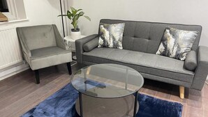 Una sala de estar con un sofá gris, un sillón gris y una mesa de centro de vidrio sobre una alfombra azul.