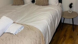 Una cama bien hecha con una manta color beige y almohadas blancas, una pequeña mesa redonda con un florero y suelo de madera.