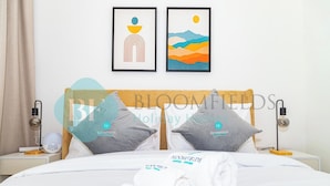 A hotel room with a neatly made bed, two pillows with the hotel's logo, a bedside table with a lamp, and two framed artworks on the wall. - Bloomfields Spacious 2BR Duplex in Oasis Masdar (Abu Dabi)