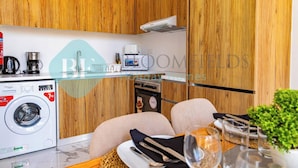 A compact kitchen with wooden cabinets, a washing machine, a coffee maker, and a toaster. - Bloomfields Spacious 2BR Duplex in Oasis Masdar (Abu Dabi)